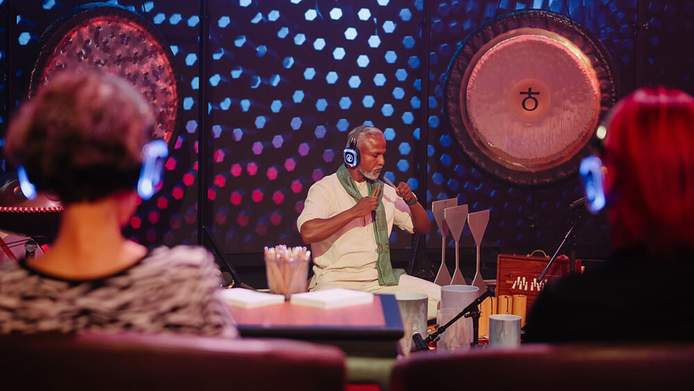 musician with gray hair, goatee playing music onlookers with headphones listen