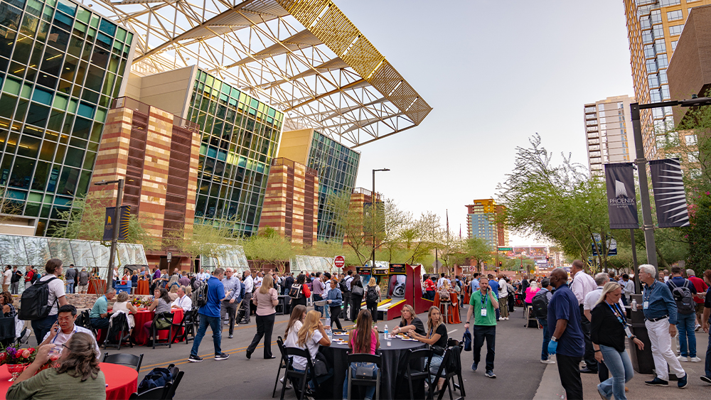 Exterior view of the Phoenix Convention Center, showcasing its modern architectural design with glass panels, geometric structures, and a vibrant urban setting under a clear sky. A large group of people some walking, other participating in activities, and sitting at tables.