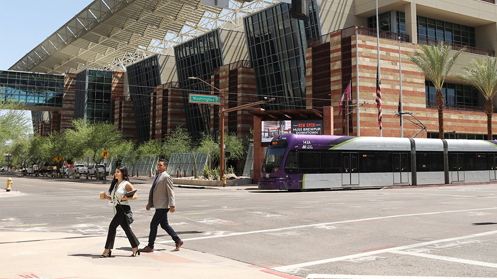 Street-level view of the Phoenix Convention Center with a light rail train passing in front, palm trees lining the street, and two pedestrians crossing at a sunny intersection.