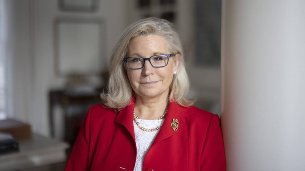 woman with glasses, red suit jacket leaning againsst white wall