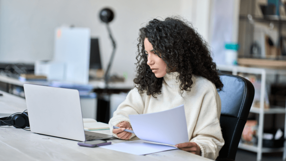 Young Latin business woman checking bills on a computer, holding paper